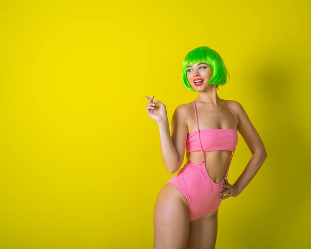 Beautiful woman in a green short wig and pink bikini posing on a yellow background. Portrait of a girl with sensual red lips. - 写真・画像