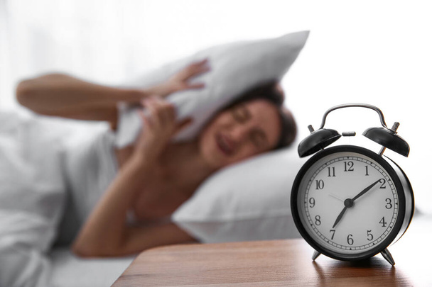 Young woman covering ears with pillow at home in morning, focus on alarm clock - 写真・画像