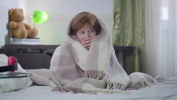 Portrait of cute little Caucasian boy sitting on bed covered with blanket and looking around. Scared child with red hair left home alone. Childhood, loneliness, fear. - Video