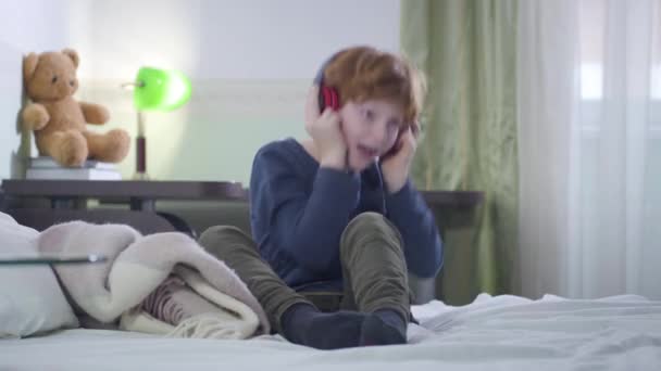 Portrait of cheerful Caucasian little boy holding headphones with hands and singing. Redhead child listening to rock music in earphones. Leisure, lifestyle, childhood. - Video, Çekim