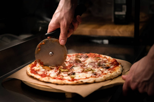 Chef cutting freshly baked delicious pizza with pizza cutter on professional kitchen. Close up of a man cutting pizza on wooden board. Traditional italian food restaurant. Low key, hand's close up - Zdjęcie, obraz