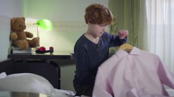 Portrait of cheerful Caucasian little boy taking pink shirt and grimacing. Joyful kid choosing outfit in his room. Childhood, lifestyle, fashion. - Materiał filmowy, wideo