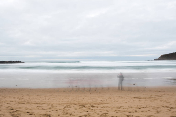 Lunga esposizione di una spiaggia con persone di passaggio e con surfisti in acqua
 - Foto, immagini