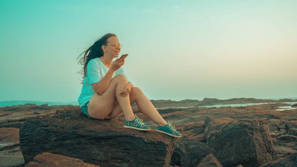 石の上に座って海辺のスマートフォンで音声メッセージを録音するリラックスした女性。楽しい女の子のサイドビュー休暇を楽しんで携帯電話でストーニーショアで話す時間を過ごす. - 写真・画像