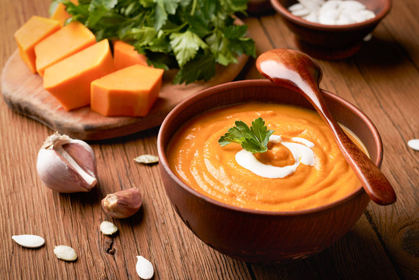 Closeup brown bowl of pumpkin soup with a wooden spoon and raw pumpkin with parsley on a cutting board - Photo, Image