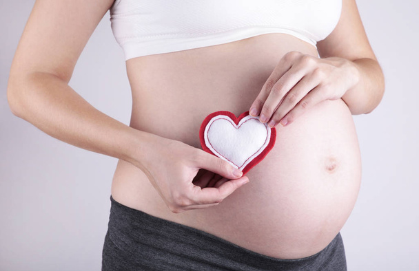Kobiety w ciąży trzymające symbol serca umieszczone na brzuchu na szarym tle - Zdjęcie, obraz
