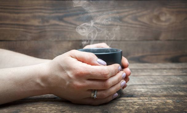 Черная чашка с кофе или чаем в руках женщины на деревянном фоне. Пар поднимается
 - Фото, изображение