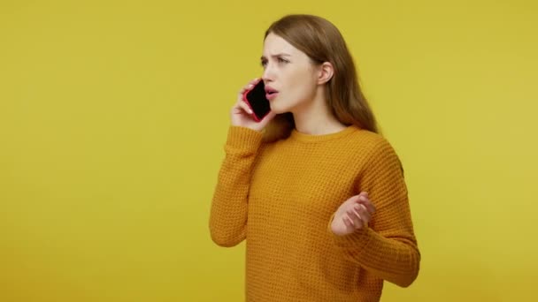 Cute girl in pullover listening carefully to conversation on her cell phone and saying wow, expressing amazement shock, discussing incredible news. indoor studio shot isolated on yellow background - Felvétel, videó