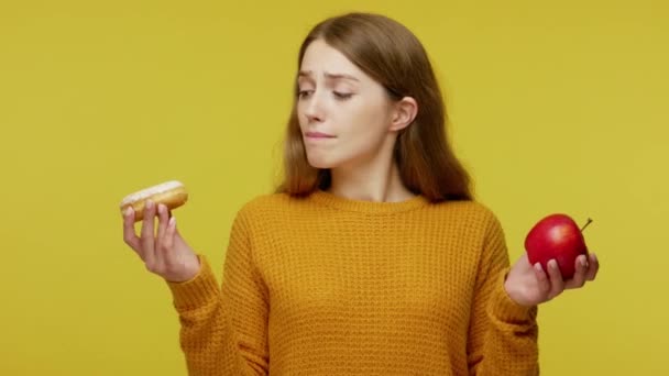 Beautiful slim girl in sweater holding apple and donut, making choice between healthy fruit and junk food, useful via harmful nutrition, diet concept. indoor studio shoot isolated on yellow background - Séquence, vidéo