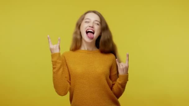 Disobedient cute girl with brown hair in pullover making rock and roll hand gesture, cool sign and showing tongue, feeling crazy carefree, naughty teenager. studio shot isolated on yellow background - Felvétel, videó
