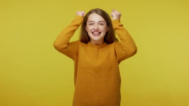 Excited champion. Enthusiastic overjoyed beautiful girl in pullover rejoicing victory, celebrating success with raised fists and dance, shouting yes. indoor studio shot isolated on yellow background - Кадри, відео