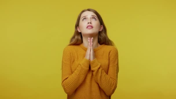 Please god, I'm begging! Cute young woman with brown hair in sweater looking up and praying with obedient imploring expression, hands holding together. indoor studio shot isolated on yellow background - Záběry, video