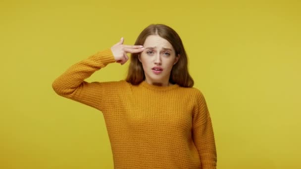Tired disappointed girl in pullover pointing finger pistol to head, feeling frustrated depressed, showing suicide gesture, shooting herself with hand gun. studio shot isolated on yellow background - Footage, Video
