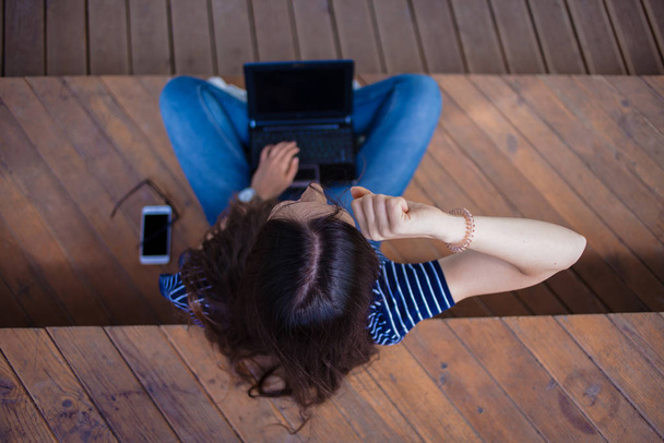 Le haut de la tête d'une étudiante brune, assise jambes croisées, sur un banc, avec un ordinateur portable ouvert, se frotte les yeux, fatiguée
. - Photo, image