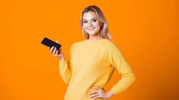 young blonde in a yellow sweater holds a smartphone in her hand with blank screen, mock up. - Фото, изображение