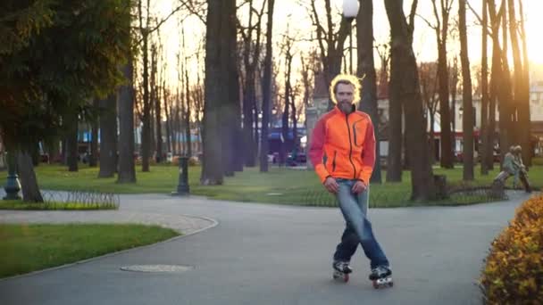 Cool bearded man wearing orange coat and jeans dances on roller skates in autumn in a central city park on a background of trees. Active sports and healthy lifestyle. Side view in slow motion. - Footage, Video