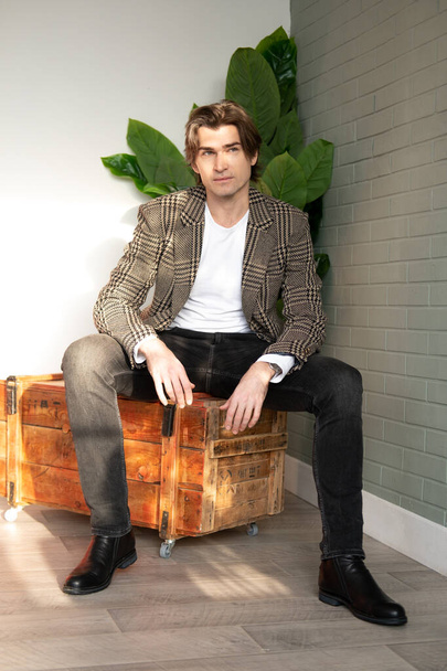 Tall handsome man sitting on the wooden box next to green plant and grey wall - Фото, зображення