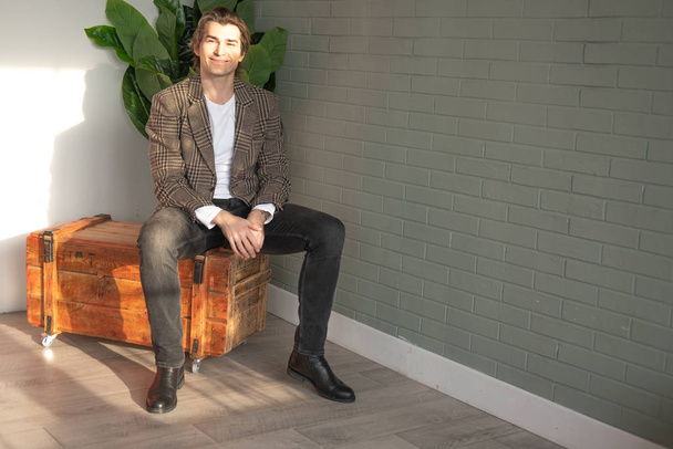 Tall handsome man sitting on the wooden box next to green plant and grey wall - Foto, Bild