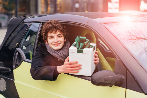 Νεαρός κατσαρομάλλης καυκάσιος αγόρασε το πρώτο μεταχειρισμένο αυτοκίνητο. Ευτυχισμένος eaxred έφηβος μαθητής κρατώντας κουτί δώρο με πράσινο τόξο κάθεται στο νέο του αυτοκίνητο. - Φωτογραφία, εικόνα