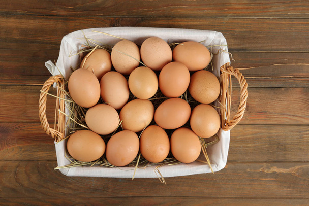 Raw chicken eggs in wicker basket on wooden table, top view - 写真・画像