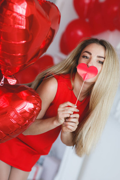 Portrait of an elegant woman in a gray room on a background of red  balloons.Attractive woman in an open, burgundy dress posing alone, standing in a set, beautiful room.Beautiful, stylish woman in a chic dress with open shoulders.Valentine's Day - Zdjęcie, obraz