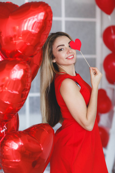 Portrait of an elegant woman in a gray room on a background of red  balloons.Attractive woman in an open, burgundy dress posing alone, standing in a set, beautiful room.Beautiful, stylish woman in a chic dress with open shoulders.Valentine's Day - Photo, image