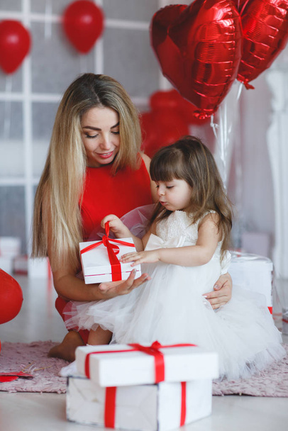 Mom and little daughter.Family portrait. Mother's day, love, family parenthood, childhood concept.Valentine's day - a young mother and little daughter in a room on the floor with gifts on the background of red balloons in the shape of a heart - Foto, Imagem