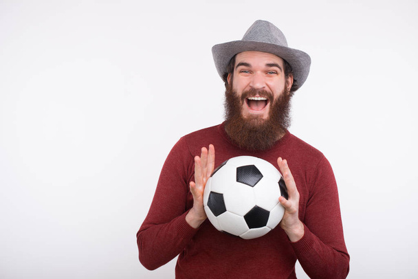 白い壁の近くにサッカーボールを両手で持っている若い幸せな髭の男. - 写真・画像
