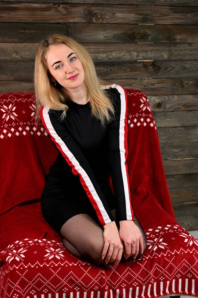 Portrait of a pretty young blonde woman with minimal makeup in a sports dress on a wooden background. Sits on a red sofa opposite the camera in various poses. - Foto, Imagem