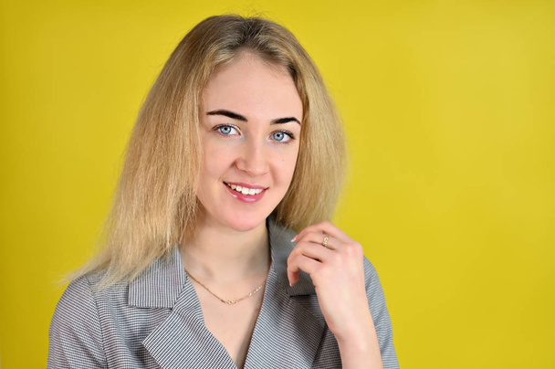 Close-up portrait of a pretty cute smiling young blonde business woman with minimal makeup in a gray suit on a yellow background. It stands directly opposite the camera in various poses. - Фото, изображение