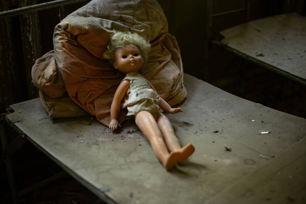 Old creepy doll on children's bed in abandoned kindergarten - Fotoğraf, Görsel