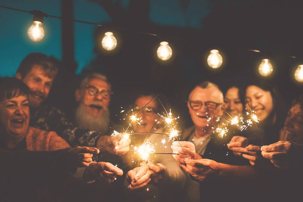 Happy family celebrating new year eve with sparkler outdoor - Group people with different ages and ethnicity having fun together in night party outside - Youth and elderly friendship lifestyle concept - Photo, Image