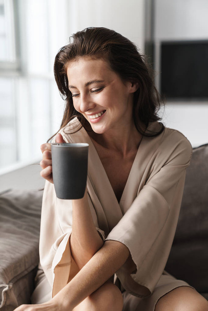 Image of young woman sitting on sofa at home and drinking tea in - Foto, imagen