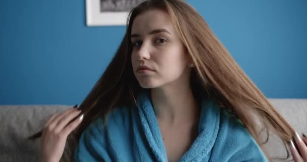 Charming woman sitting on grey couch in blue bathrobe - Záběry, video