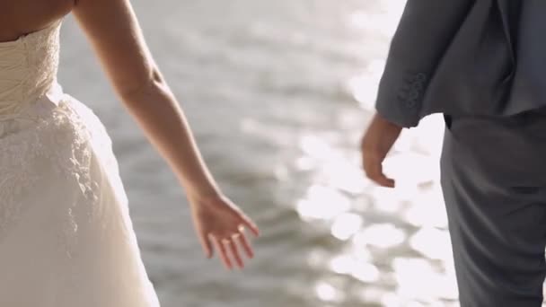 Friss házasok állnak a tenger mellett. Egymás kezét nyújtják. Vőlegény és menyasszony - Felvétel, videó