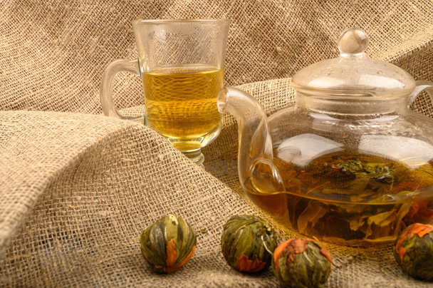 Blütentee gebrüht in einer gläsernen Teekanne, ein Glas Tee und Bällchen Blütentee auf einem Hintergrund aus grobem selbstgesponnenem Stoff. Nahaufnahme. - Foto, Bild