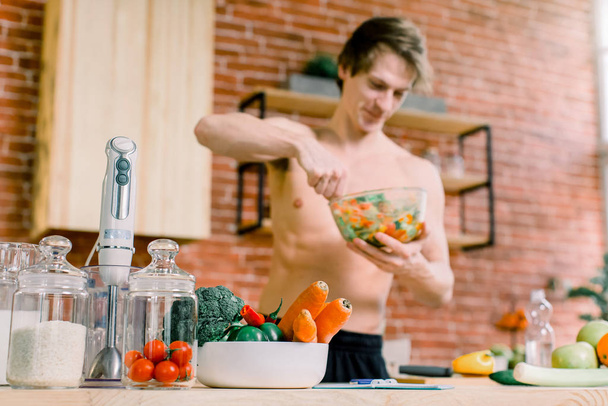 Its so delicious. Casual happy young man preparing salad at home in loft kitchen and smiling. - 写真・画像