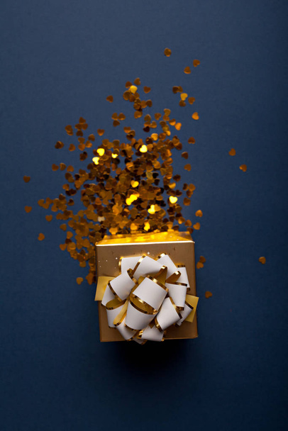 Golden gift box with bow over classic blue color backgroundwith many shiny gold hearts sparkles - Fotó, kép