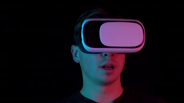 Молодой человек в очках VR крупным планом. Человек, погруженный в виртуальную реальность, делает движения руками. Синий и красный свет падает на человека на черном фоне
. - Кадры, видео