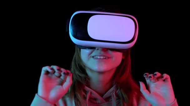Nuori nainen VR-laseissa lähikuvassa. Virtuaalitodellisuuteen upotettu nainen tekee liikkeitä käsillään. Sininen ja punainen valo osuu naiselle mustalla taustalla
. - Materiaali, video