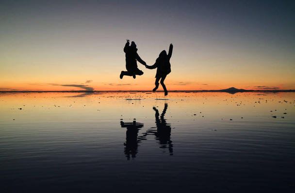 Silueta de pareja feliz saltando sobre el increíble efecto espejo de Uyuni Salt Flats contra el cielo del atardecer, Bolivia, América del Sur
 - Foto, Imagen