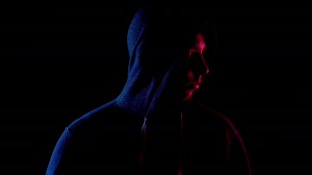 A young man in a hood looks around. An attacker is standing in the dark. Blue and red light falls on a person on a black background. - Video, Çekim