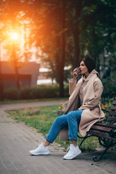 Όμορφη νεαρή γυναίκα κάθεται σε ένα παγκάκι πίνοντας καφέ απολαμβάνοντας στο πάρκο - Φωτογραφία, εικόνα