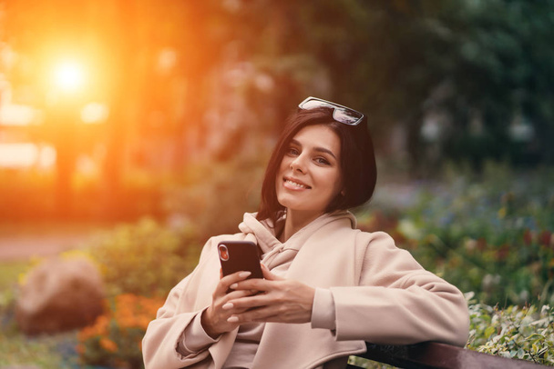 Счастливая девушка с помощью смартфона в городском парке сидит на скамейке - Фото, изображение