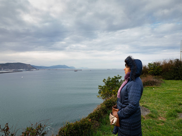 Kobieta w płaszczu patrząc na morze i port Bilbao w oddali, z wiatrem. - Zdjęcie, obraz