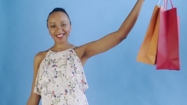 African american woman is posing with shopping bags on blue Background in Studio. Happy Woman Holding Shopping Colorful Bags. white dress with flowers - Filmati, video