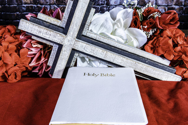 Λευκή Βίβλος σε κόκκινο δίπλα σε ξύλινο σταυρό με κόκκινα και λευκά λουλούδια στο παρασκήνιο - Φωτογραφία, εικόνα