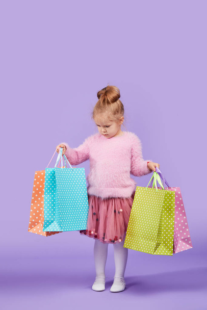 Portrait of a happy little child girl standing on a purple background holding shopping bags, package. Looking at the camera. - Фото, зображення