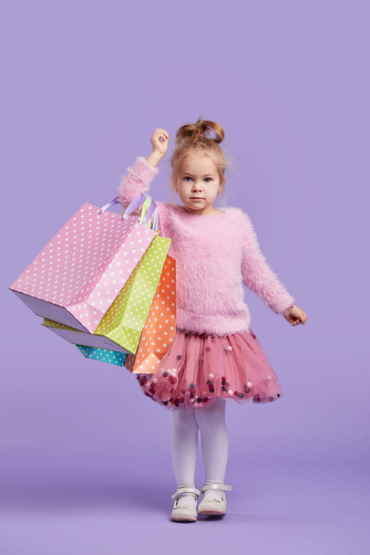Portrait of a happy little child girl standing on a purple background holding shopping bags, package. Looking at the camera. - Φωτογραφία, εικόνα