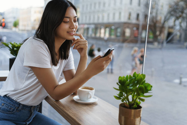 Leisure, weekends and urban lifestyle concept. Carefree young modern girl using smartphone, scroll social media as sitting alone cafe, lean coffee table, drink cappucino smiling, look mobile display - Φωτογραφία, εικόνα